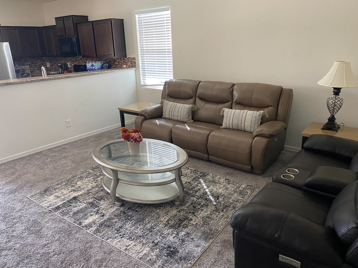 A welcoming living area features two comfortable seating options, including a brown leather couch and a reclining sofa. A glass-top coffee table sits on a patterned area rug, complemented by natural light filtering in from a nearby window. A kitchenette is visible in the background.