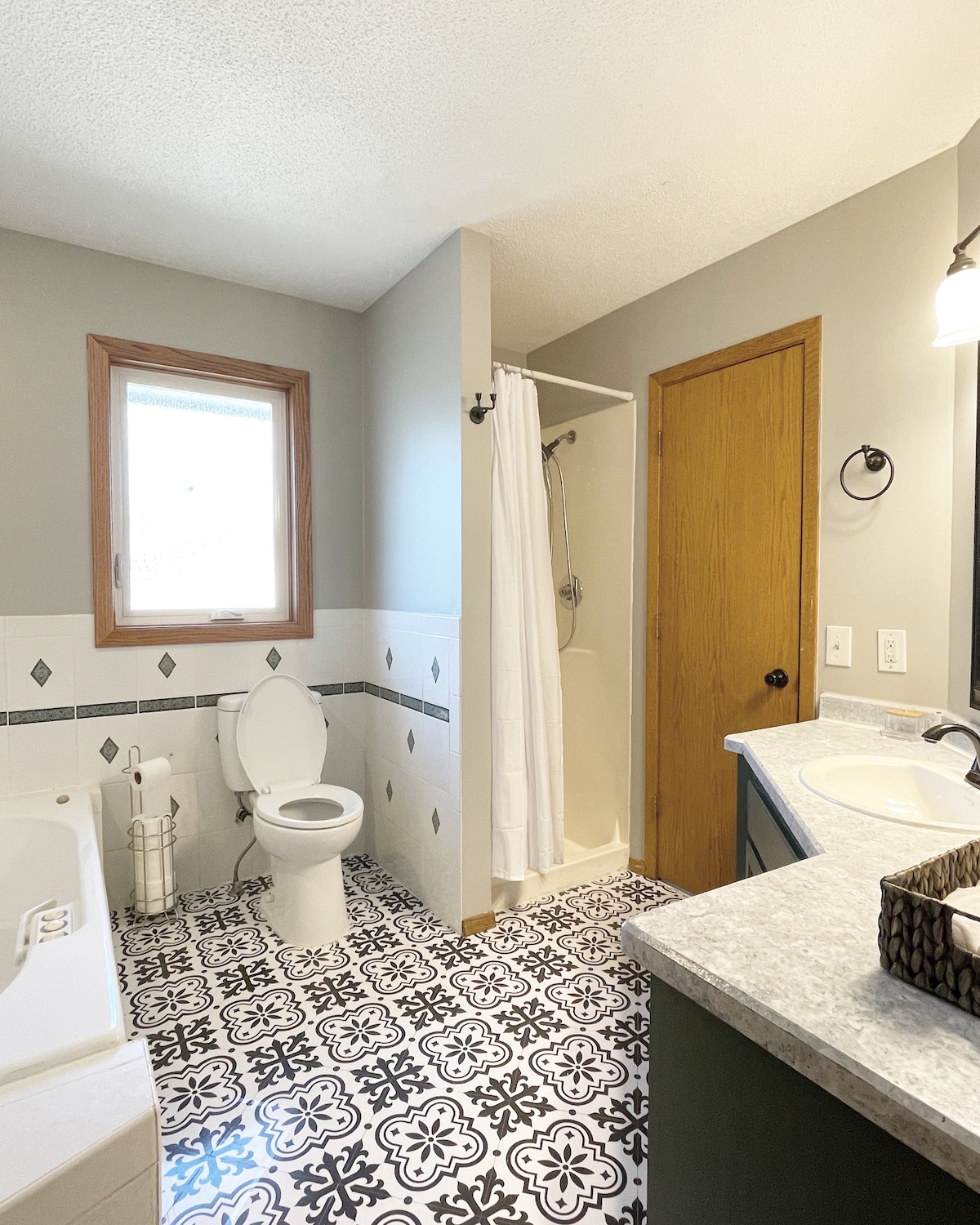 A bright bathroom is displayed, featuring a spacious vanity with a stone countertop and a basket for toiletries. A bathtub and separate shower stall are included, with a white curtain concealing the shower area. Decorative black and white tiles cover the floor, adding character to the space.