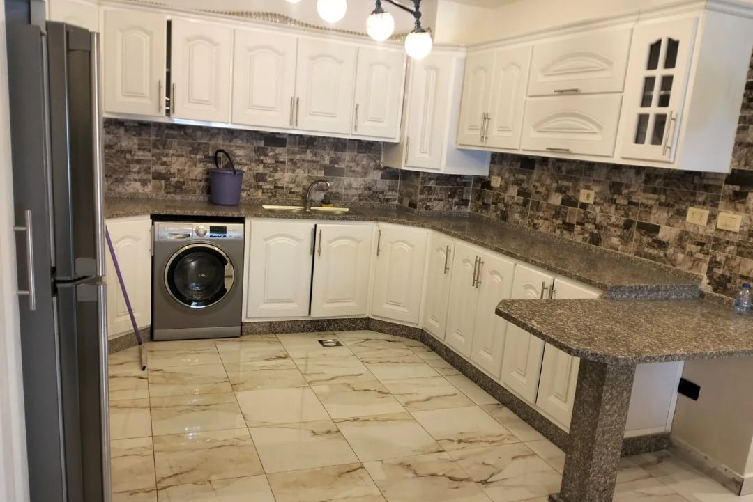 A modern kitchen features white cabinetry and a spacious countertop with a textured stone finish. Stainless steel appliances, including a washing machine, are integrated seamlessly. Neutral-colored tiles create an even floor space, while decorative lighting fixtures hang above the area.