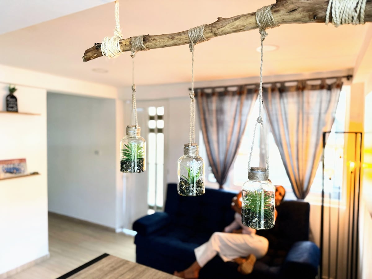 Suspended glass jars containing small plants are displayed from a wooden branch, adding a touch of greenery to the space. A dark blue sofa is visible in the background, with sheer curtains framing a window that allows natural light to fill the room.