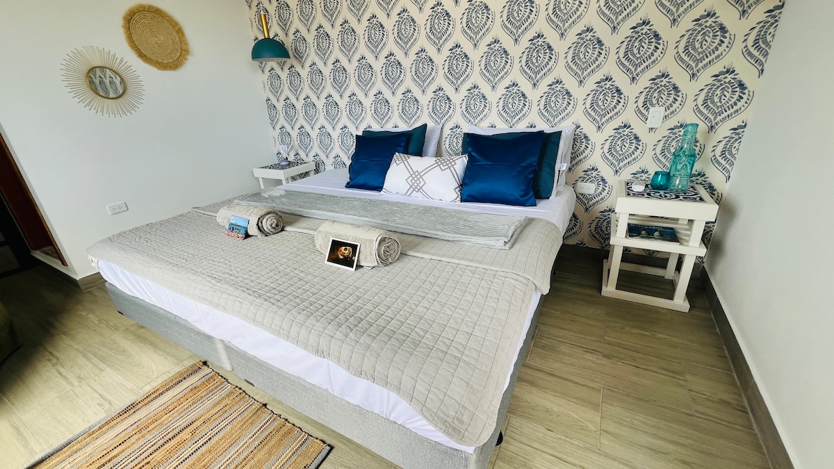 A bright sleeping area features a spacious bed dressed with a light grey quilt and decorative pillows in blue tones. The patterned wall adds visual interest. A bedside table is positioned on either side, and decorative items provide a welcoming touch.