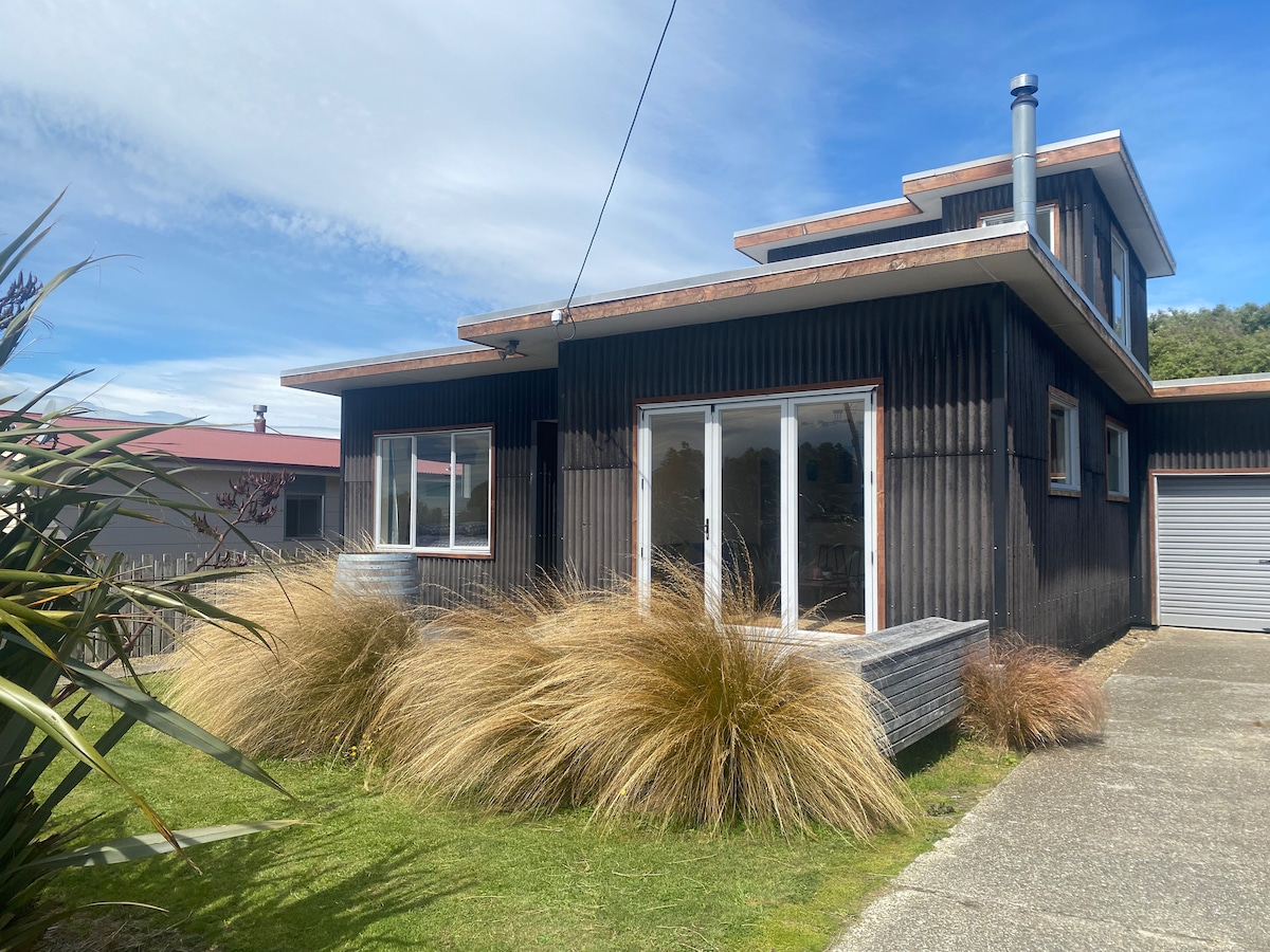 The exterior of a two-story home features dark wooden cladding and a spacious deck. Large glass doors provide access to the interior, allowing natural light to fill the space. Lush grass and native vegetation are present in the garden area, contributing to the tranquil setting.