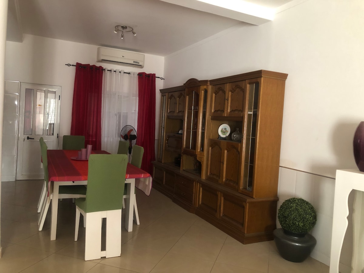 A dining area is presented with a long table surrounded by green chairs. A warm wooden cabinet displays decorative items along the wall. Light streams in through a window, while sheer curtains add a soft touch to the space.