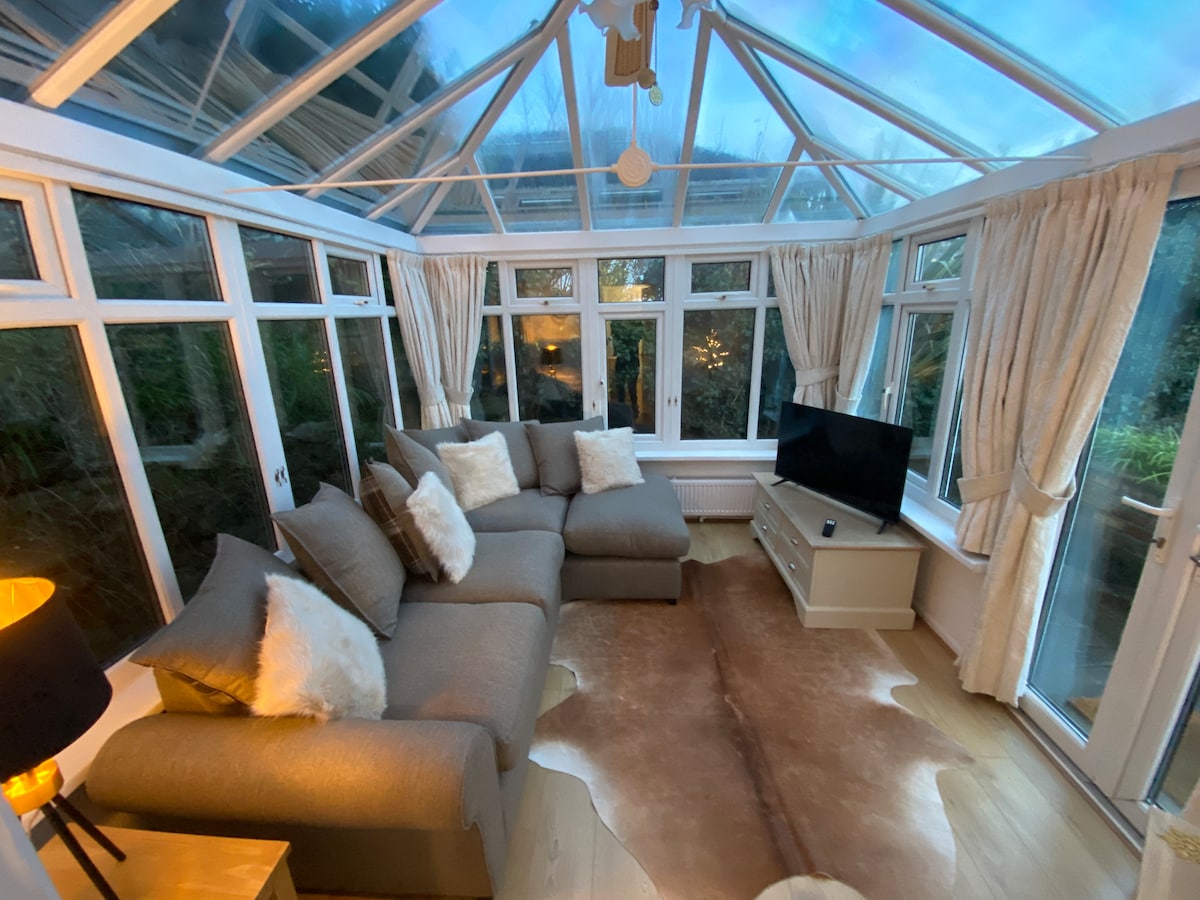 The conservatory features a comfortable sectional sofa adorned with soft pillows, accompanied by a television mounted on a light-colored console. Natural light floods the space through numerous glass panels, creating a bright and inviting atmosphere, enhanced by subtle decor elements.