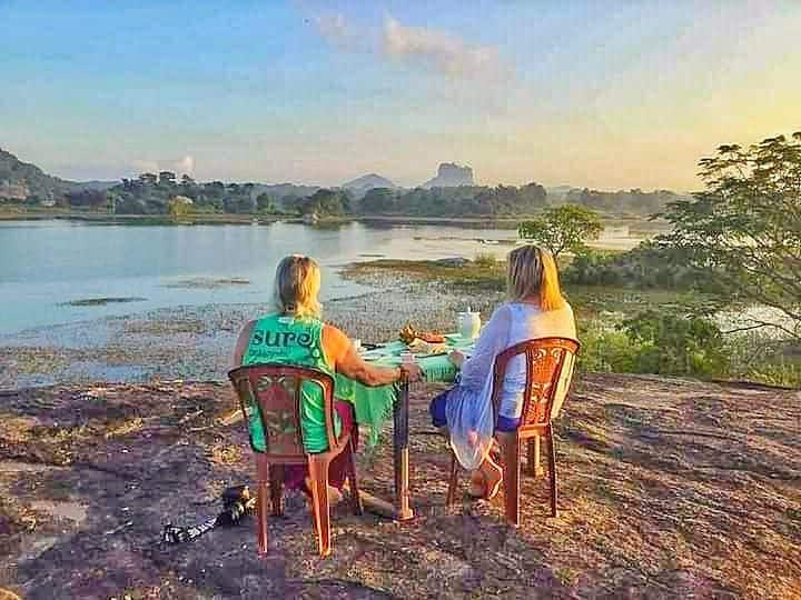 Two guests are seated at a small table on a rocky outcrop overlooking a tranquil lake, with lush greenery surrounding the area. Scenic views of distant hills and Sigiriya Lion Rock are visible in the background, complemented by a soft golden light from the setting sun.