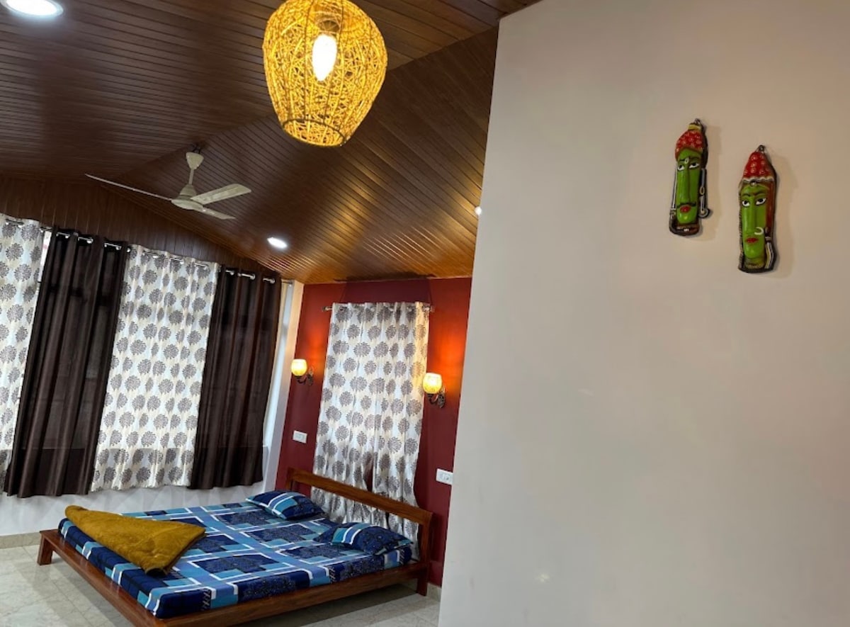 A spacious sleeping area features a wooden bed adorned with a patterned blue and white bedspread. Decorative wall masks add character to the room, which is illuminated by a woven pendant light. Large windows are dressed with patterned curtains and dark shades.