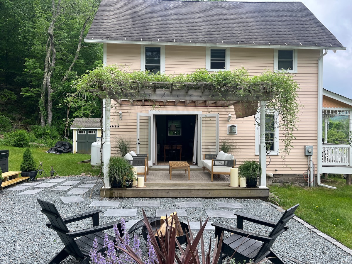 The exterior of the cottage is revealed, showcasing a welcoming entrance with a covered porch adorned with greenery. A stone pathway leads to a seating area, surrounded by mature plants and a fire pit, creating an inviting outdoor space.