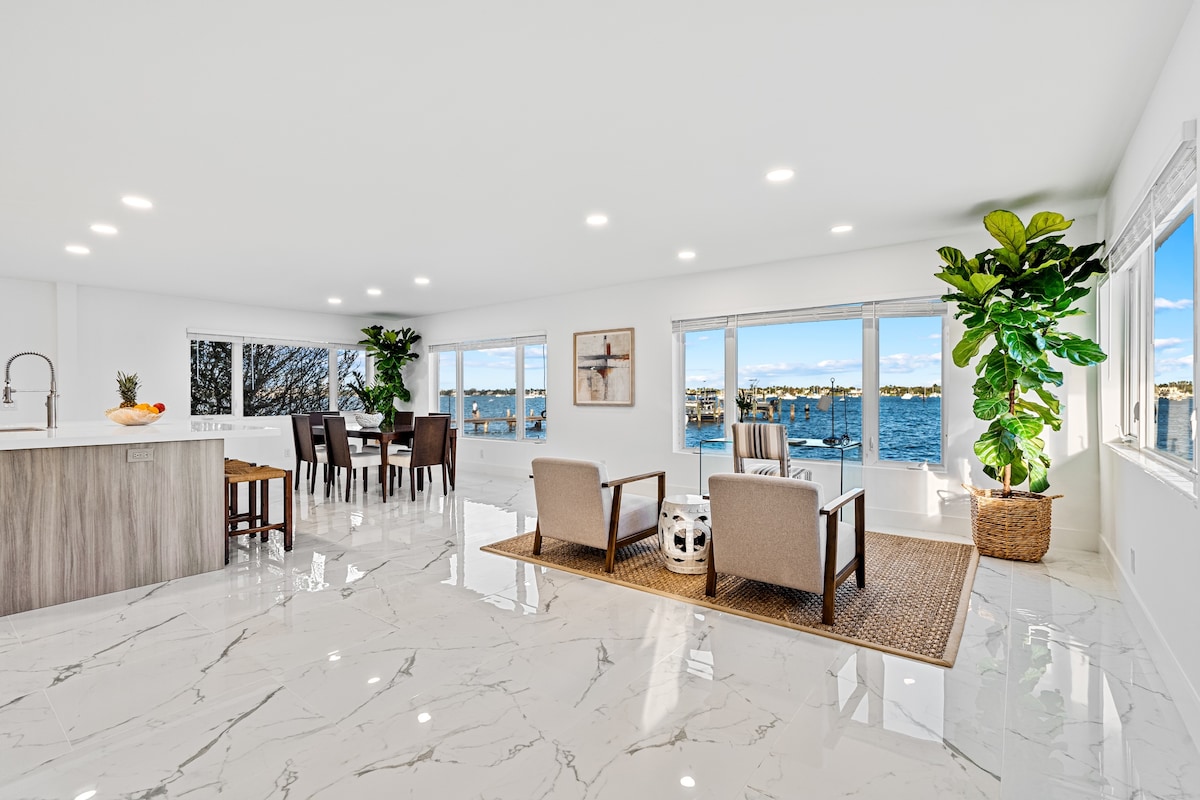 The open living area features modern furniture arranged to encourage conversation, with large windows offering views of the intracoastal waterway. A spacious kitchen island is present, and indoor plants add a touch of greenery to the bright, airy space.
