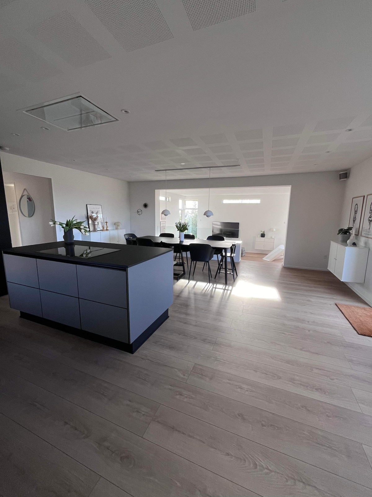 A modern kitchen area is showcased, featuring a spacious kitchen island with a dark countertop. Surrounding dining chairs are arranged around a rectangular table, while ample natural light fills the room through large windows. The open layout connects to a relaxing living space.