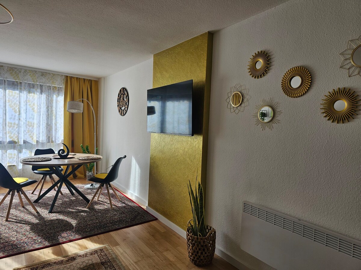 A dining area is centered around a circular table accompanied by four chairs. A television is mounted on the wall, and decorative mirrors are arranged in a stylish pattern. A potted plant adds a touch of greenery, while natural light filters through sheer curtains.