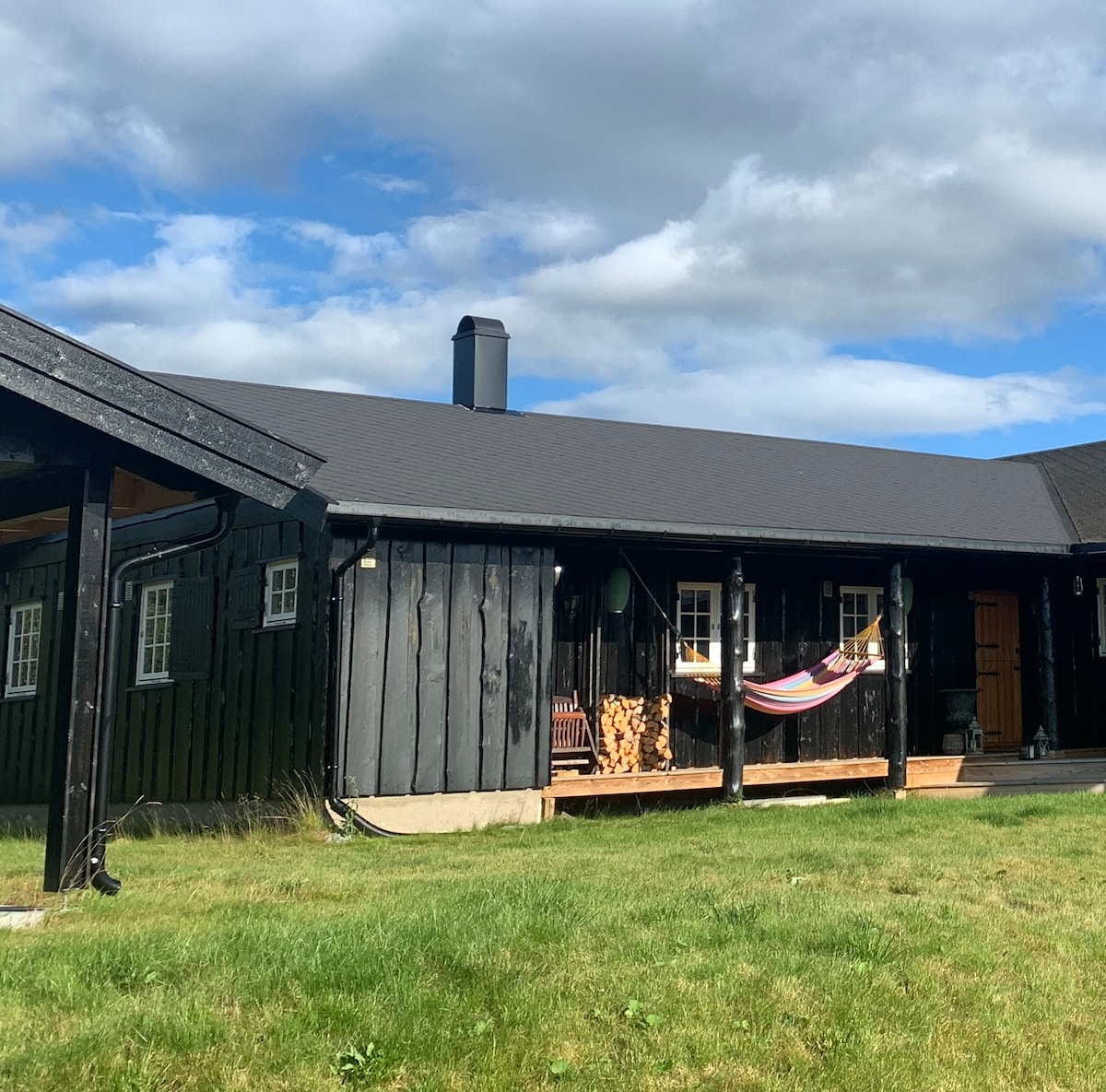 The exterior of a black wooden cabin is depicted, showcasing a spacious porch with a colorful hammock. A stack of firewood is neatly arranged beside the entrance, and expansive grass areas surround the cabin, complemented by a partly cloudy sky.