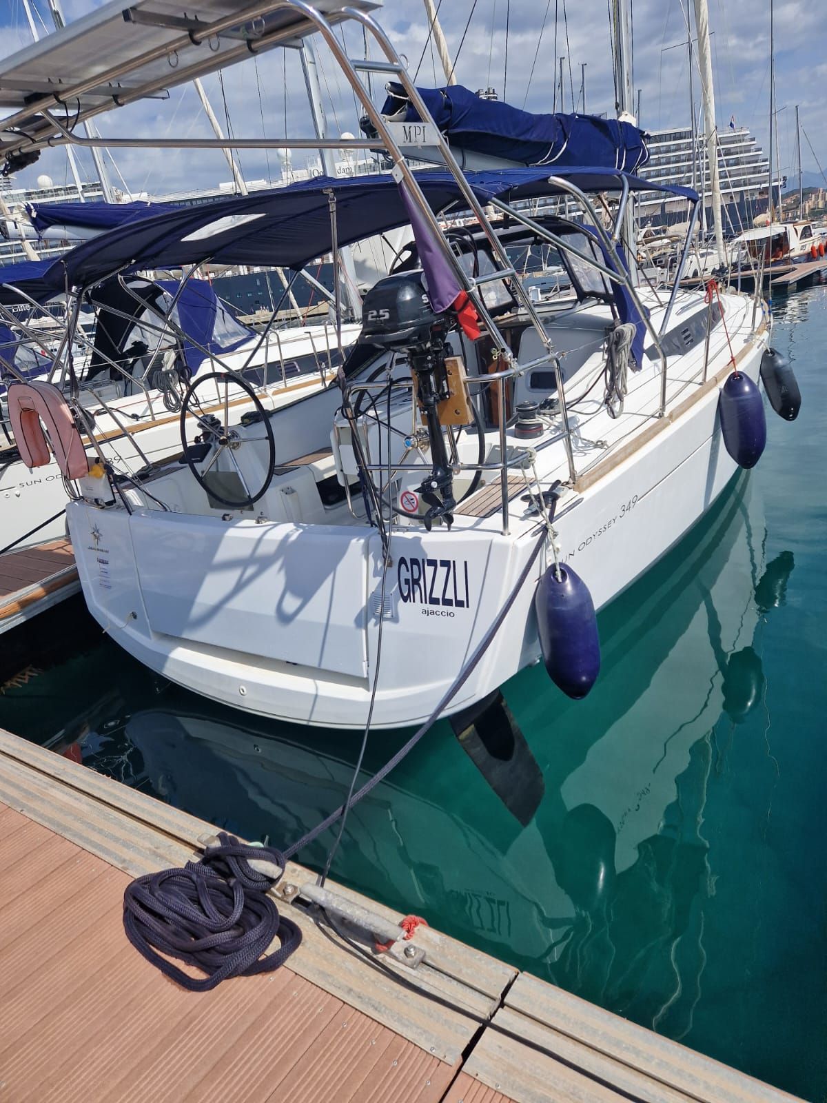 A sailboat is docked at a marina, featuring a white hull and blue accents. The vessel is equipped with a canopy and various sailing equipment. Reflections of the boat can be seen in the calm water, surrounded by additional boats and marina structures in the background.