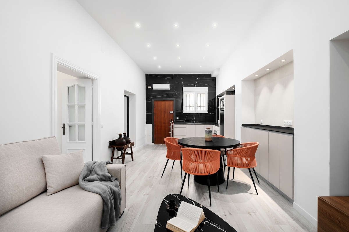 A spacious living area is visible, featuring a comfortable light-colored sofa and a round dining table surrounded by vibrant orange chairs. The sleek kitchen can be seen in the background, with modern appliances and a black accent wall. Light-colored wood flooring completes the contemporary look.