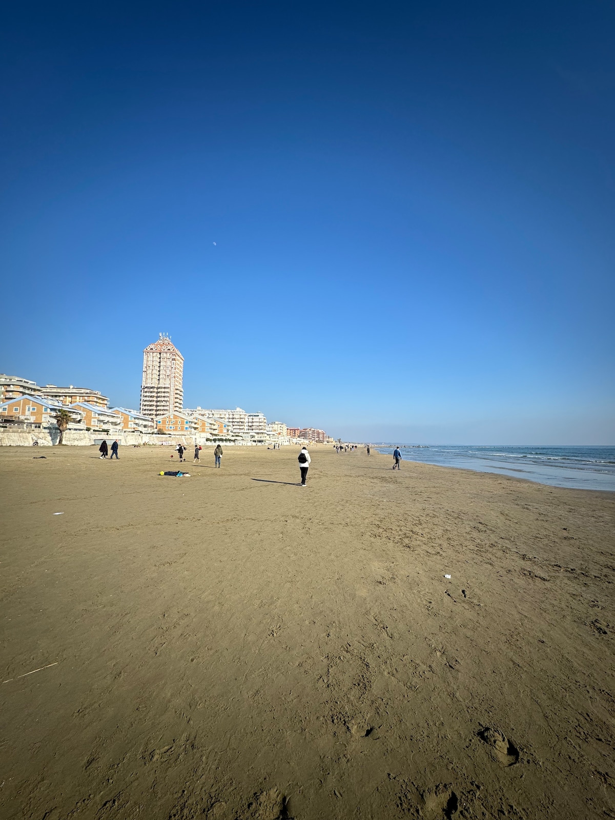 A wide sandy beach stretches alongside a calm sea under a clear blue sky. In the distance, tall buildings and residential structures are visible, while several people enjoy the beach, some walking along the shore and others engaging in leisurely activities.