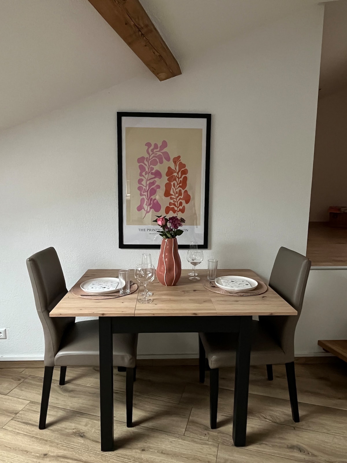 A compact dining area is shown with a wooden table set for two. Light-colored plates, glasses, and cutlery are positioned on a tabletop adorned with a decorative vase containing flowers. A framed piece of art in warm tones serves as a backdrop.