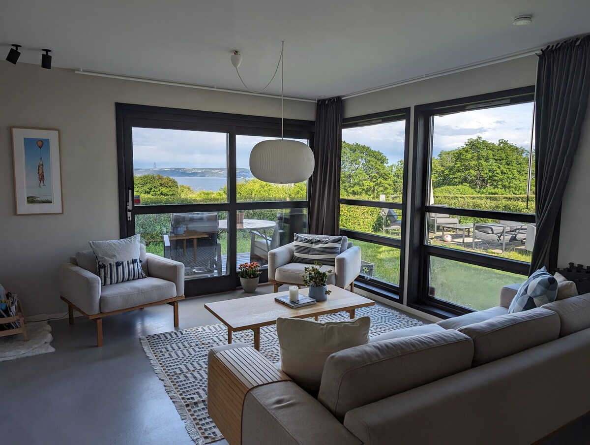 A bright living area is framed by large windows offering views of the surrounding greenery. Comfortable seating includes a spacious sofa and two armchairs, arranged around a central coffee table. Soft textiles and a light rug add warmth to the contemporary design.