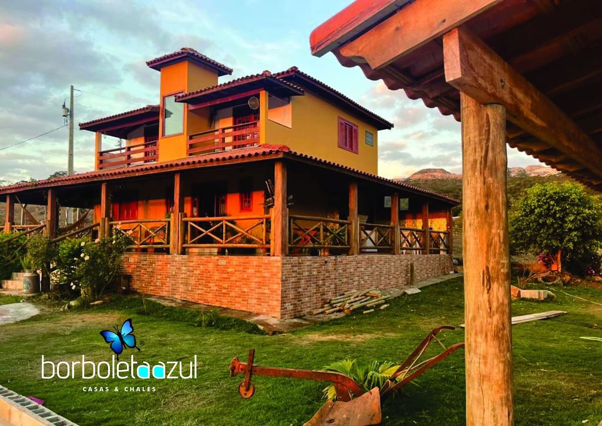 The exterior of a two-story house is displayed, featuring vibrant yellow and red tones. A large wooden deck with railings surrounds the structure, providing ample outdoor space. Lush green grass leads to a gently sloping landscape, framed by distant hills.