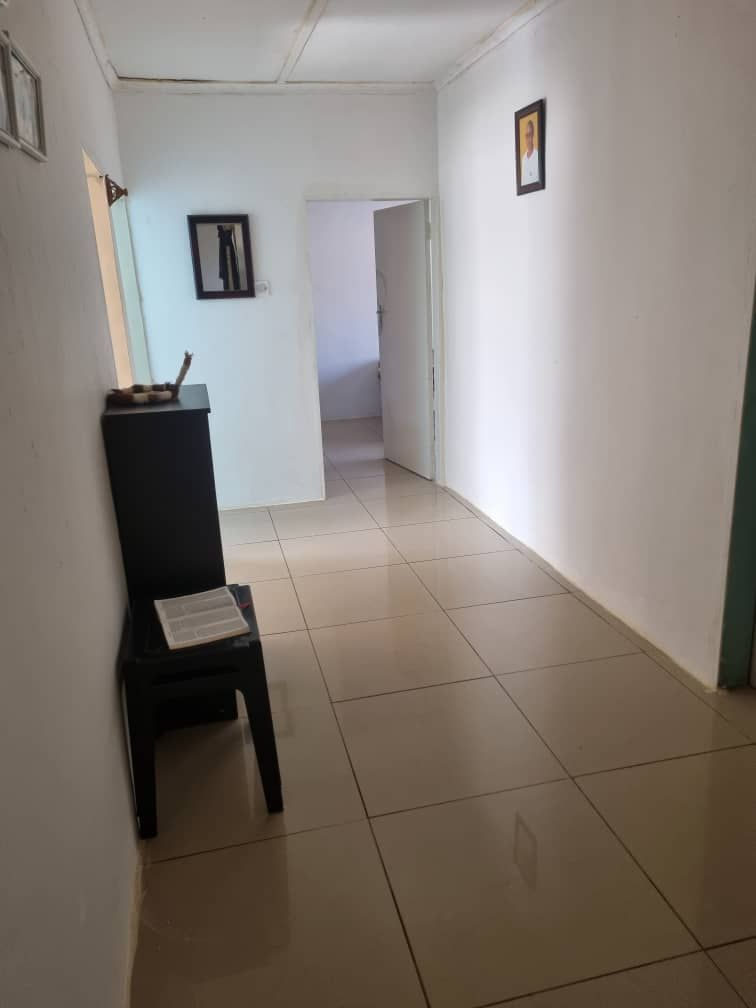 A narrow hallway features smooth, tiled flooring with a neutral tone. Light-colored walls create an airy feel, while a small black table stands against one side. Two framed pictures are mounted on the wall, leading to an open door at the end.