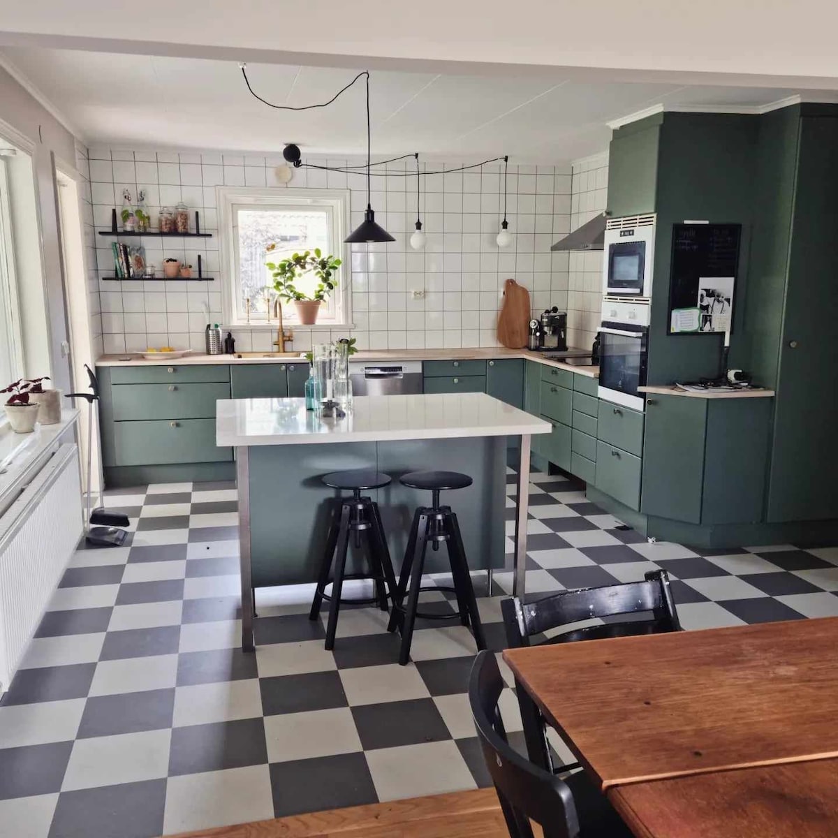 A well-equipped kitchen features a central island with two stools and a combination of green cabinetry and white backsplash tiles. The floor showcases a checkered pattern in black and white, while natural light illuminates the space through the window above the sink.