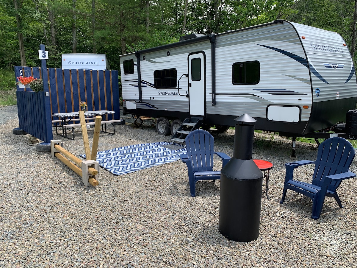 A travel trailer is parked on a gravel area surrounded by greenery. An outdoor seating setup includes two blue chairs and a black fire pit. A patterned rug lies on the ground, adding comfort to the space. A wooden fence with a sign identifies the site.