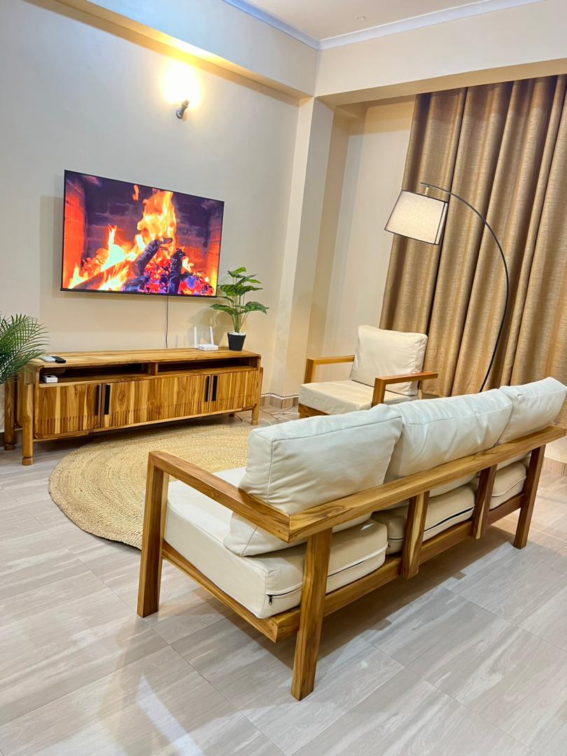 A cozy living area features a wooden sofa with cream-colored cushions, accompanied by a matching armchair. A low wooden console holds a television displaying a fireplace. Soft golden curtains frame the space, complemented by a large area rug and a potted plant.