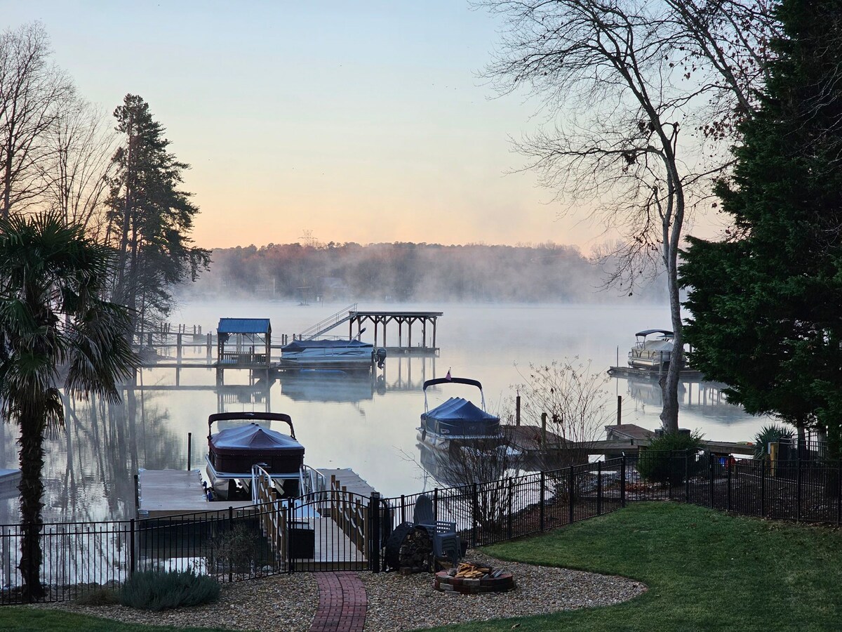 A serene lakeside view shows calm water enveloped in a gentle mist at dawn. Several boats are docked, and a wooden pier extends into the water. Lush greenery and trees frame the scene, complemented by structured landscaping leading to the water's edge.