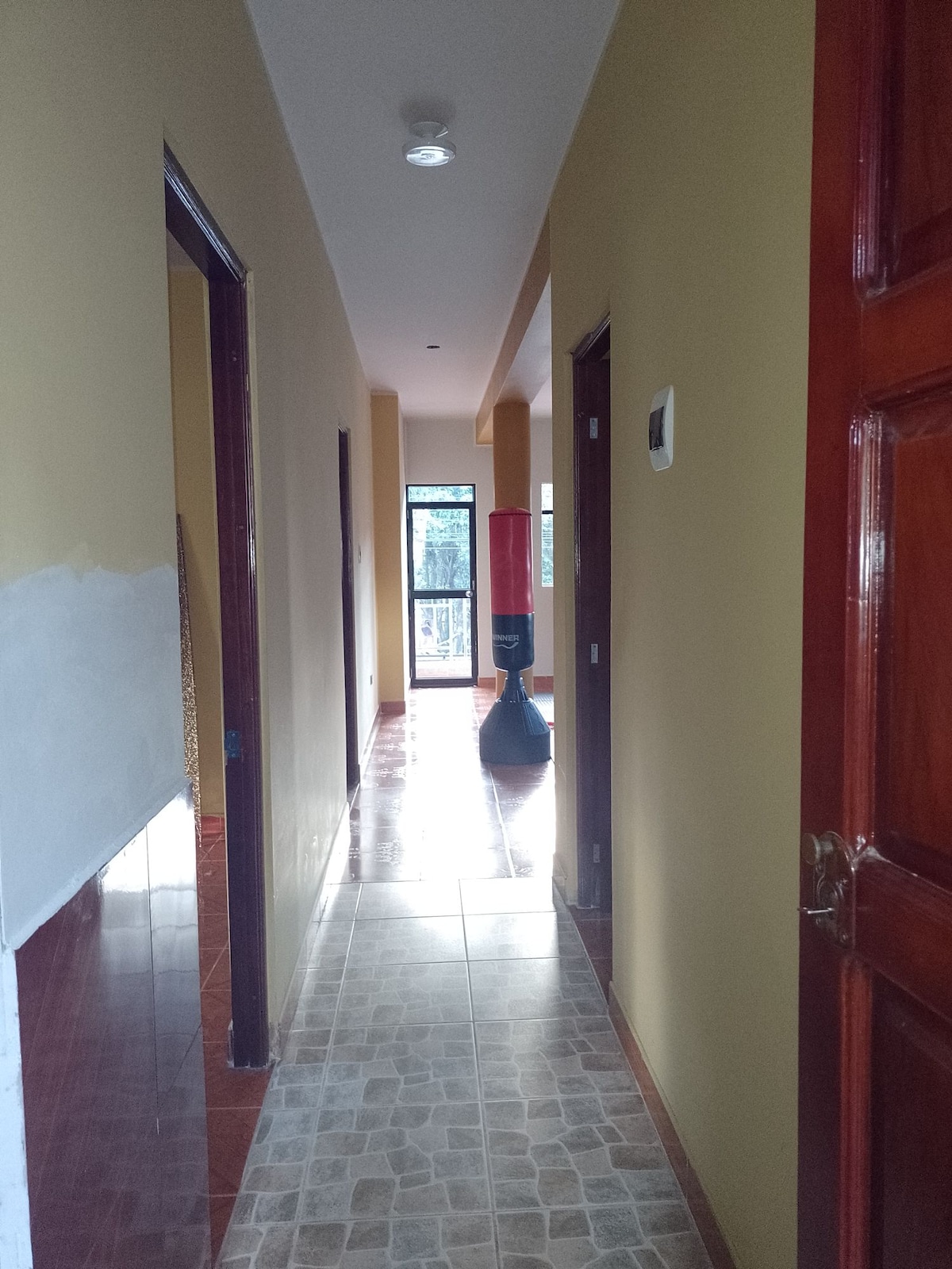 A hallway is visible, lined with wooden doors on both sides. Natural light is seen coming from a large window at the end of the corridor, illuminating the tiled floor. A punching bag is positioned against the wall, adding a unique element to the space.