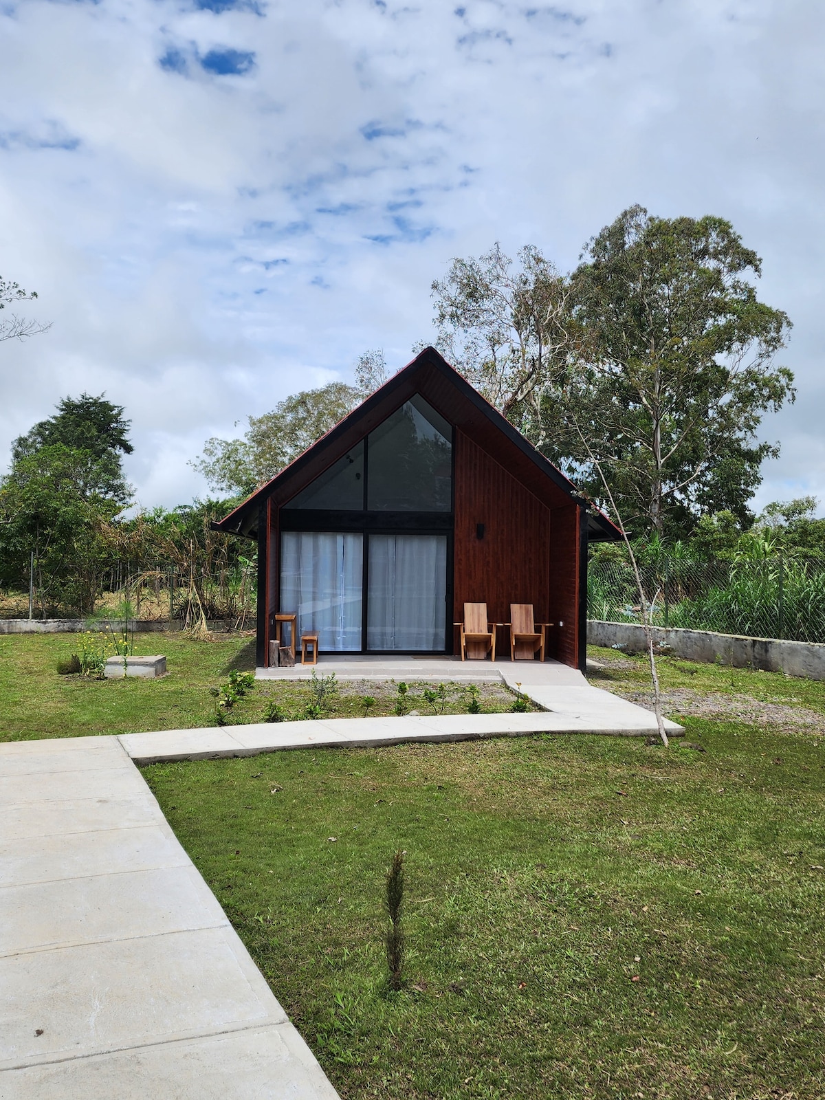 The exterior of a contemporary cabin is shown, featuring a sloped roof and large glass doors that open to a small terrace. Two wooden chairs are placed on the terrace, surrounded by a well-maintained green lawn and landscaped areas. The cabin is set against a backdrop of trees and a cloudy sky.