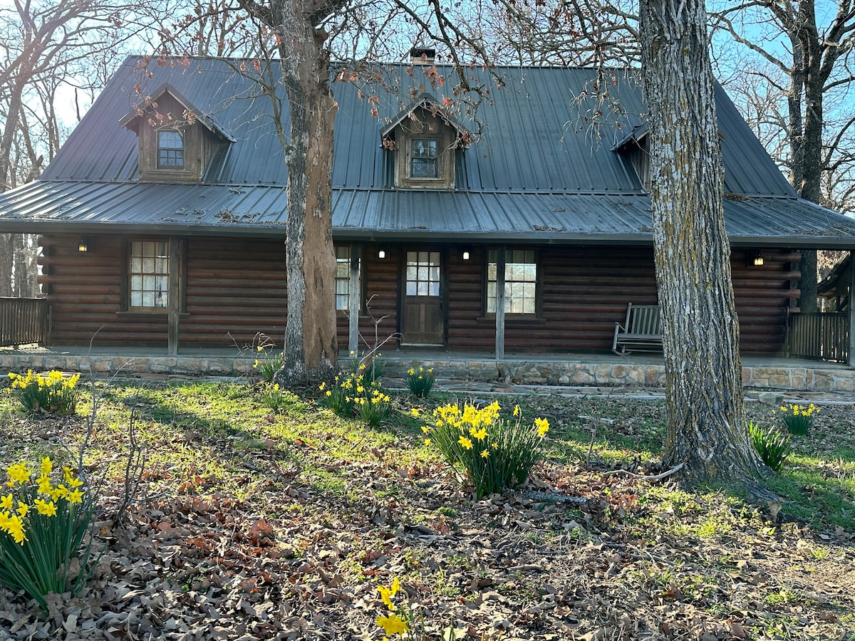 A rustic log cabin features a charming metal roof and multiple windows, allowing natural light to fill the interior. Yellow flowers bloom in the foreground, complementing the wooden architecture. The porch includes a swing, enhancing the inviting outdoor space.