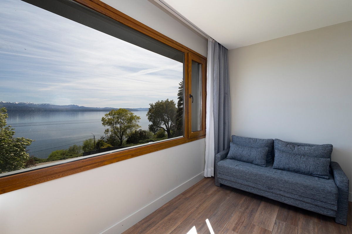 A cozy seating area features a grey couch positioned near a large window, allowing for unobstructed views of Lake Nahuel Huapi. Natural light floods the space, highlighting the warm tones of the wooden window frame and the soft texture of the couch.