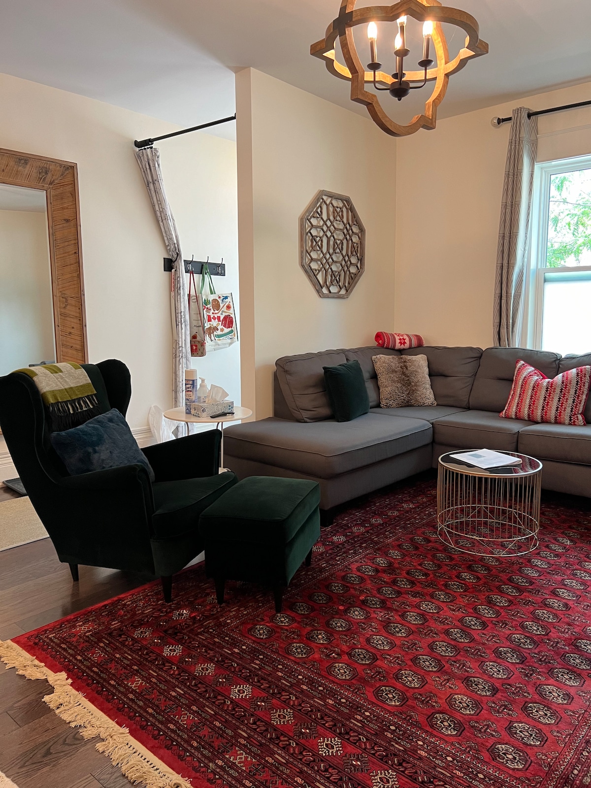 A comfortable living area is highlighted by a gray sectional sofa and a cozy armchair in dark green. A decorative area rug with a rich pattern covers the floor. Natural light filters through a nearby window, complemented by simple curtains and a stylish chandelier overhead.