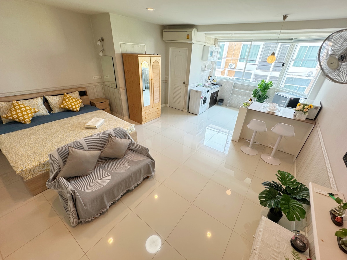 A spacious room includes a comfortable bed with decorative pillows and a cozy seating area. A dining table with chairs and essential appliances, such as a washing machine, are visible. Large windows provide ample natural light, enhancing the room's open layout.