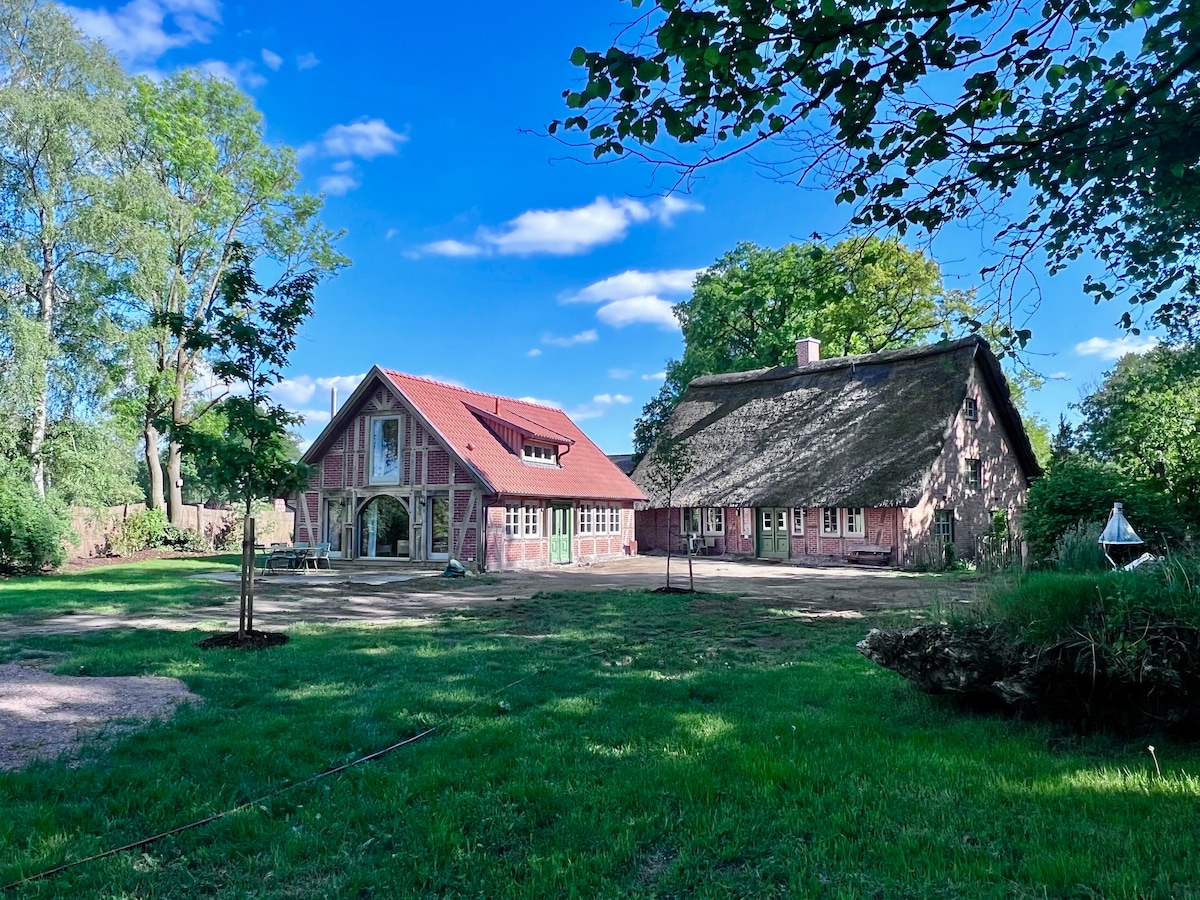 The image shows an inviting holiday cottage with a thatched roof alongside a beautifully restored building made of traditional brick. Lush green grass surrounds the structures, and trees provide dappled shade, creating a serene outdoor setting. A clear blue sky fills the background.