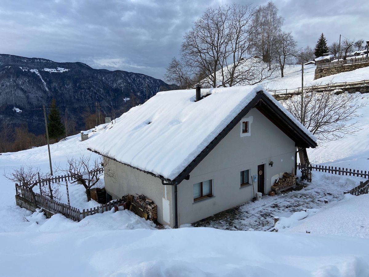 A charming house is nestled in a snowy landscape, surrounded by a wooden fence. A sloped roof is blanketed with snow, while a small stack of firewood is visible at the side. Rolling mountains and trees complement the serene setting.