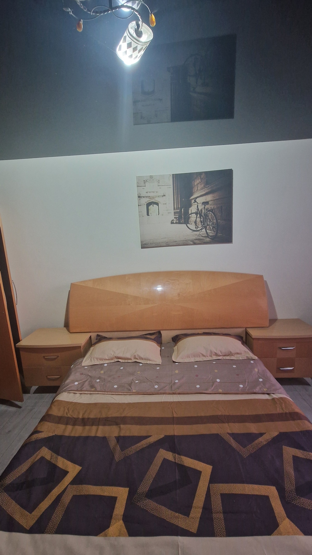 A bedroom featuring a spacious bed with neutral-colored bedding and two decorative pillows. Nightstands are located on either side, and a simple light fixture illuminates the room. Two framed photographs hang above the bed, adding a touch of design to the space.