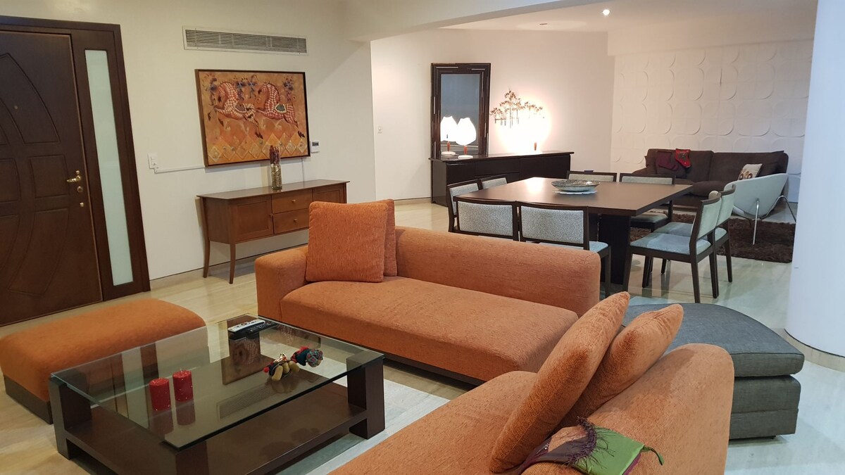 An open living area features a large orange sectional sofa surrounding a glass coffee table. A wooden dining table sits nearby, accompanied by several chairs. Art is displayed on the wall, complementing the bright space illuminated by soft overhead lighting.