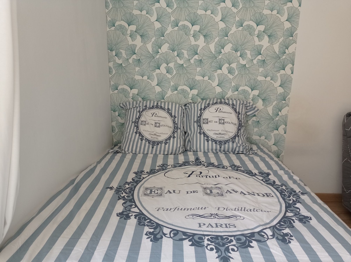 A spacious bed is adorned with striped bedding featuring a lavender scent theme. Two matching pillows with decorative prints rest against the wall, which is decorated with a soft floral pattern. Natural light filters gently through nearby curtains.