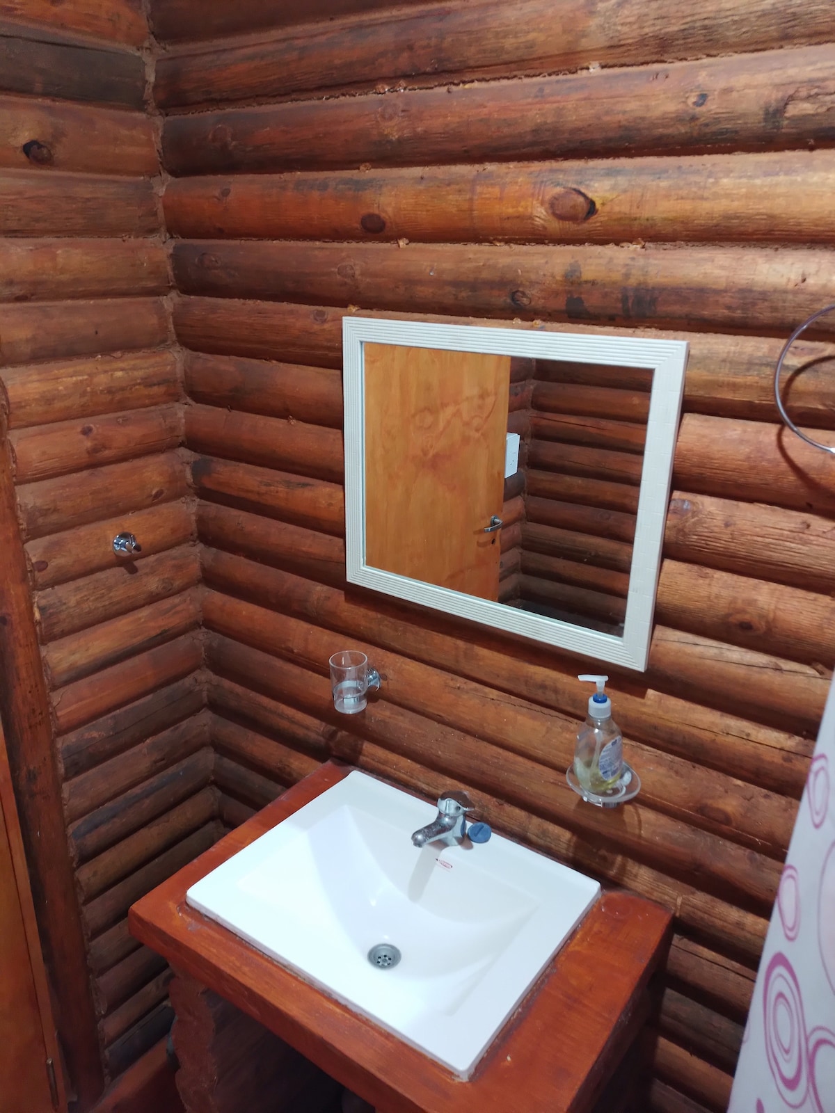 A rustic bathroom space is presented with natural wooden walls and a simple white sink. A mirror framed in white reflects the warm tones of the logs, while a soap dispenser and a glass for toiletries are mounted nearby. Soft lighting enhances the cozy ambiance.