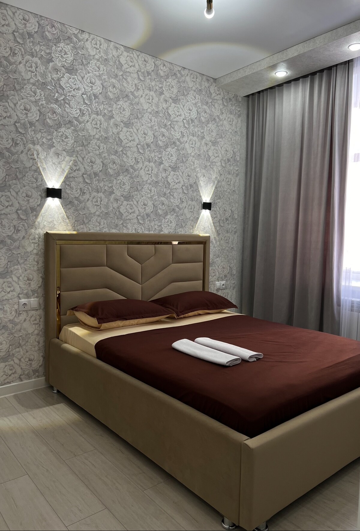 A bedroom is displayed with a stylish design featuring a spacious bed dressed in rich brown linens. Elegant wall sconces provide soft lighting alongside the window, which is dressed with light gray curtains. The patterned wallpaper adds a sophisticated touch to the space.