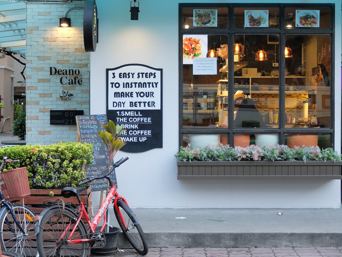 The café exterior features a welcoming entrance with large windows showcasing baked goods and a cozy interior. A sign with steps to a better day is displayed prominently. Outdoor seating is available, along with a bicycle parked nearby, enhancing the local atmosphere.