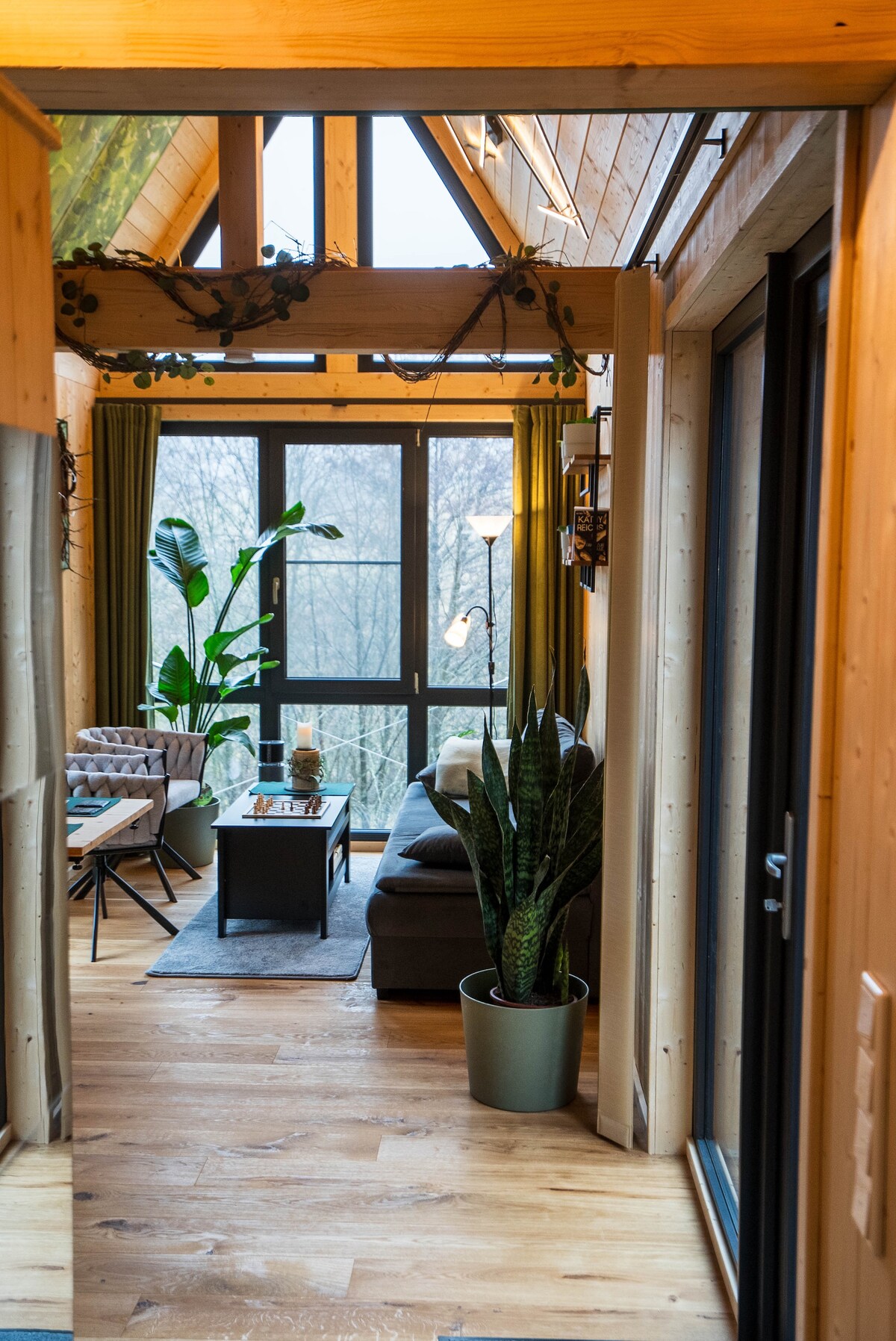 A hallway leads to a cozy living area featuring a comfortable sofa and two armchairs. Large windows provide natural light and views of greenery outside. A coffee table sits at the center, with a potted plant nearby adding an inviting touch.