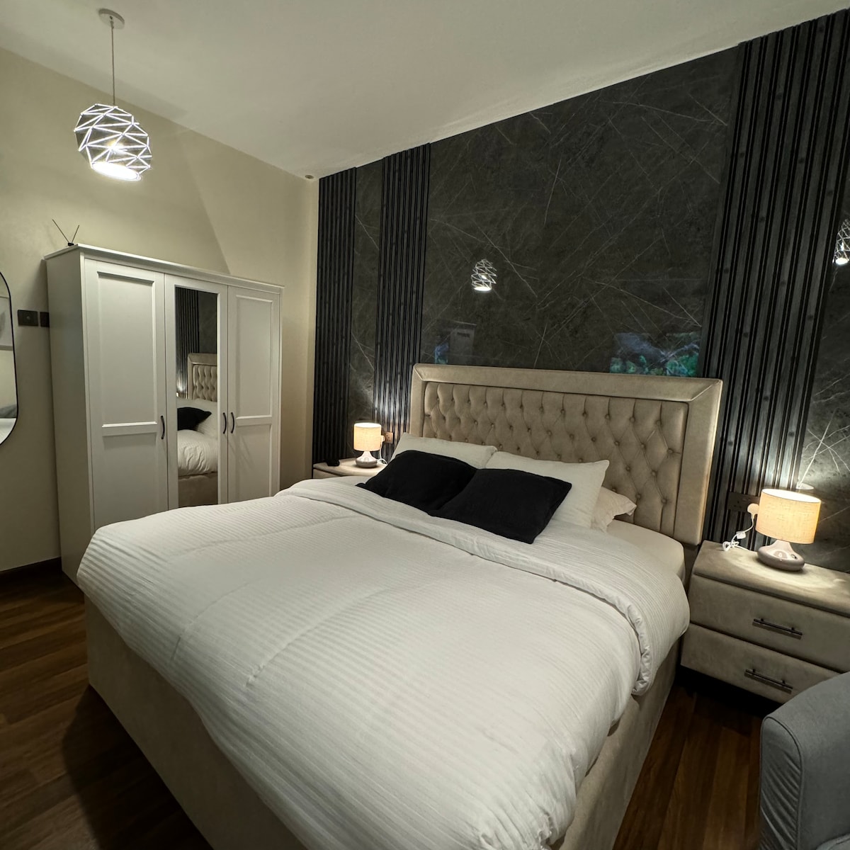 A modern bedroom features a spacious bed dressed in crisp white linens, flanked by two lamps on bedside tables. A large wardrobe resides against one wall, while a circular mirror adds depth. Textured dark panels accentuate the headboard area.