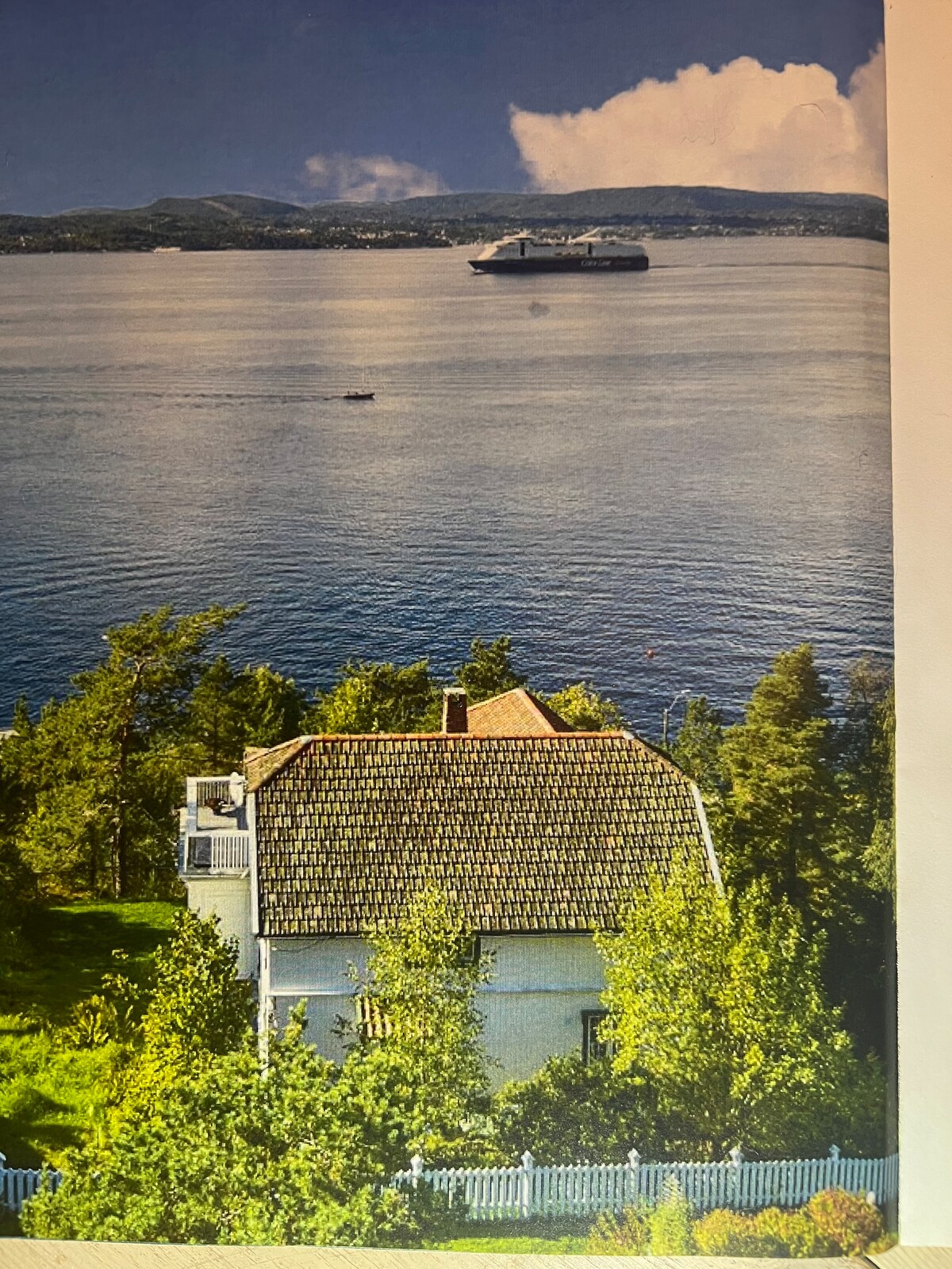 A picturesque view of the villa from above is visible. The structure, featuring a sloped roof, is surrounded by lush greenery. The serene Oslo Fjord extends beyond, with a ferry gently gliding across the water, framed by distant hills under a bright sky.