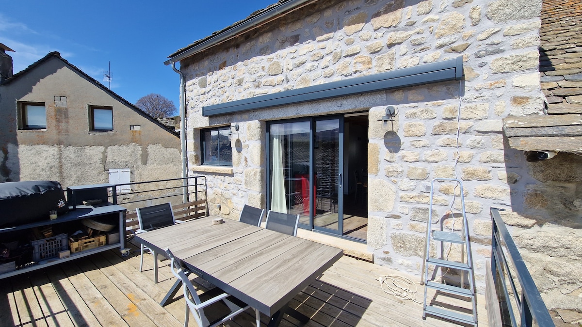 A stone exterior of the house is showcased, featuring large glass doors that open to a spacious wooden deck. A dining table with several chairs is positioned outside, while a grill is visible on one side, enhancing the outdoor living experience.