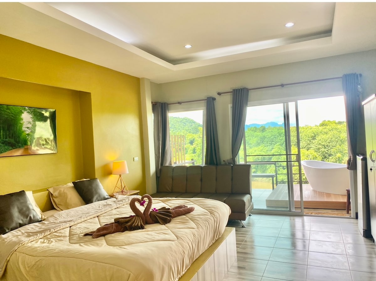 A spacious sleeping area features a large bed with neutral-toned bedding and decorative towels arranged in a heart shape. A seating area with a sleek sofa is present, and glass doors provide a view of lush greenery outside, leading to a private balcony.