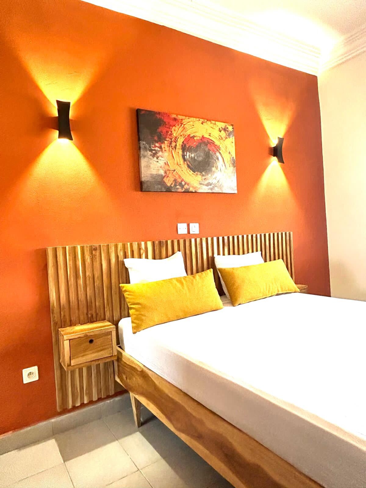 A cozy sleeping area is defined by a wooden bed frame featuring a spacious mattress. Two yellow accent pillows are placed against a backdrop of rich orange walls, highlighted by soft wall sconces. A decorative wall art piece adds a colorful touch.