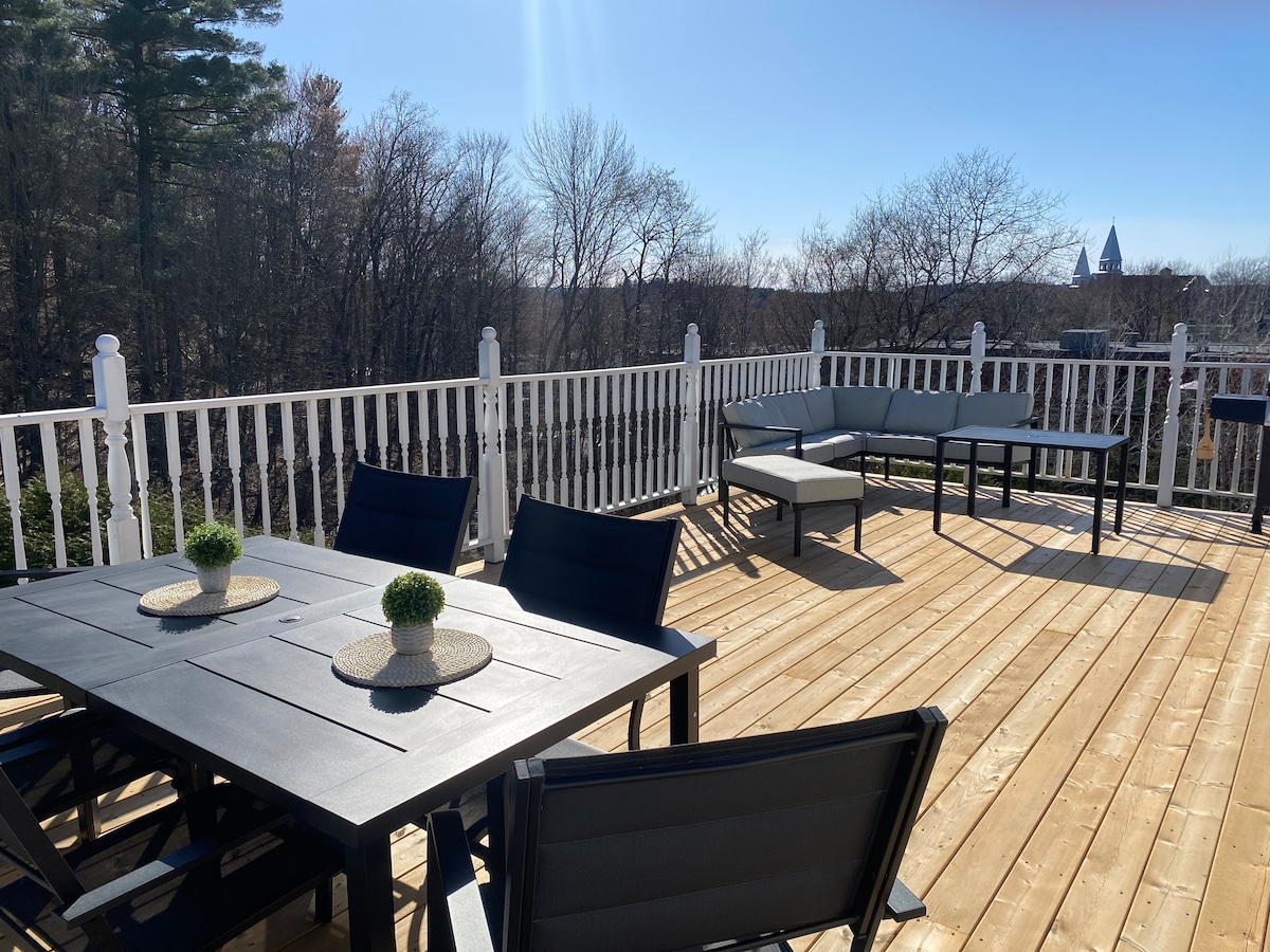 An expansive terrace features a wooden deck, furnished with a dark table and chairs. A sectional seating area offers additional comfort. The surrounding trees provide a natural backdrop, while distant structures can be seen beyond the railing, creating a sense of openness and connection to the outdoors.