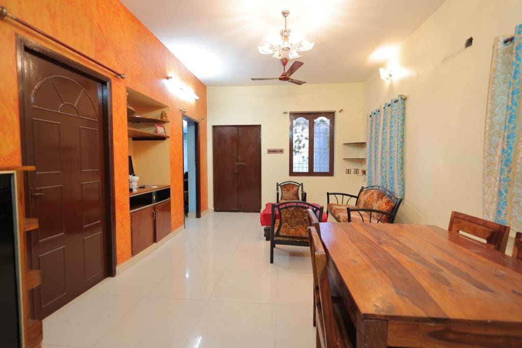 A bright living area features an orange accent wall, complemented by light-colored walls. A wooden dining table and chairs are included, along with two seating chairs beside a small shelf. Natural light filters through the windows covered with blue curtains.