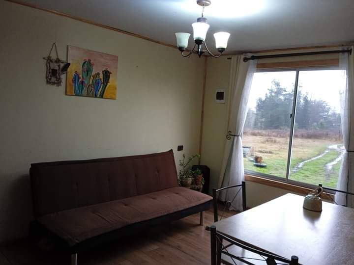 A light-filled living area features a brown futon, a small dining table with delicate black metal chairs, and a colorful artwork on the wall. Natural light enters through a large window, offering views of the outdoor landscape and a peaceful, green setting.