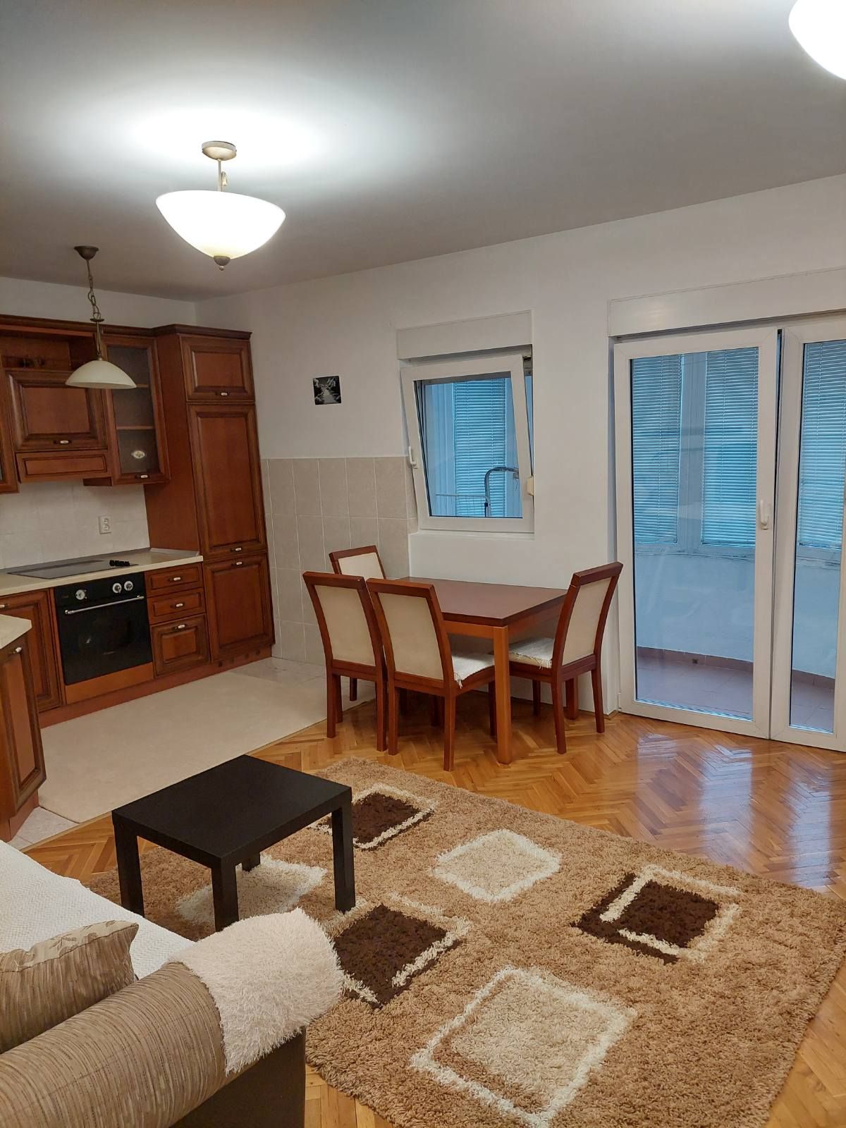 A dining area is depicted with a rectangular wooden table surrounded by four chairs. A cozy rug with geometric patterns lies on the floor. A small black coffee table is positioned nearby. The space is illuminated by a ceiling light, and large glass doors offer access to a balcony.